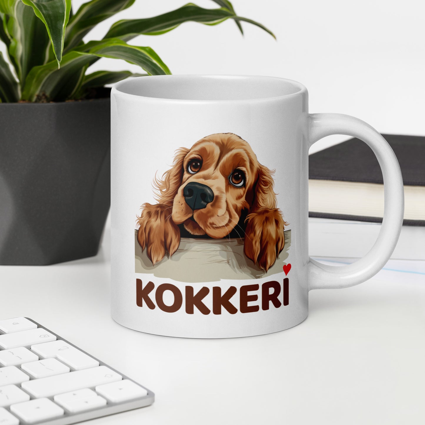 Työpöydällä valkoinen muki, jossa cockerspanielin kuva ja teksti "kokkeri" punaisella sydämellä varustettuna.