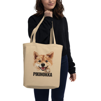 Nainen pitelee isoa beigeä kangaskassia, jossa on suomenpystykorvan kuva ja teksti "pikinokka".