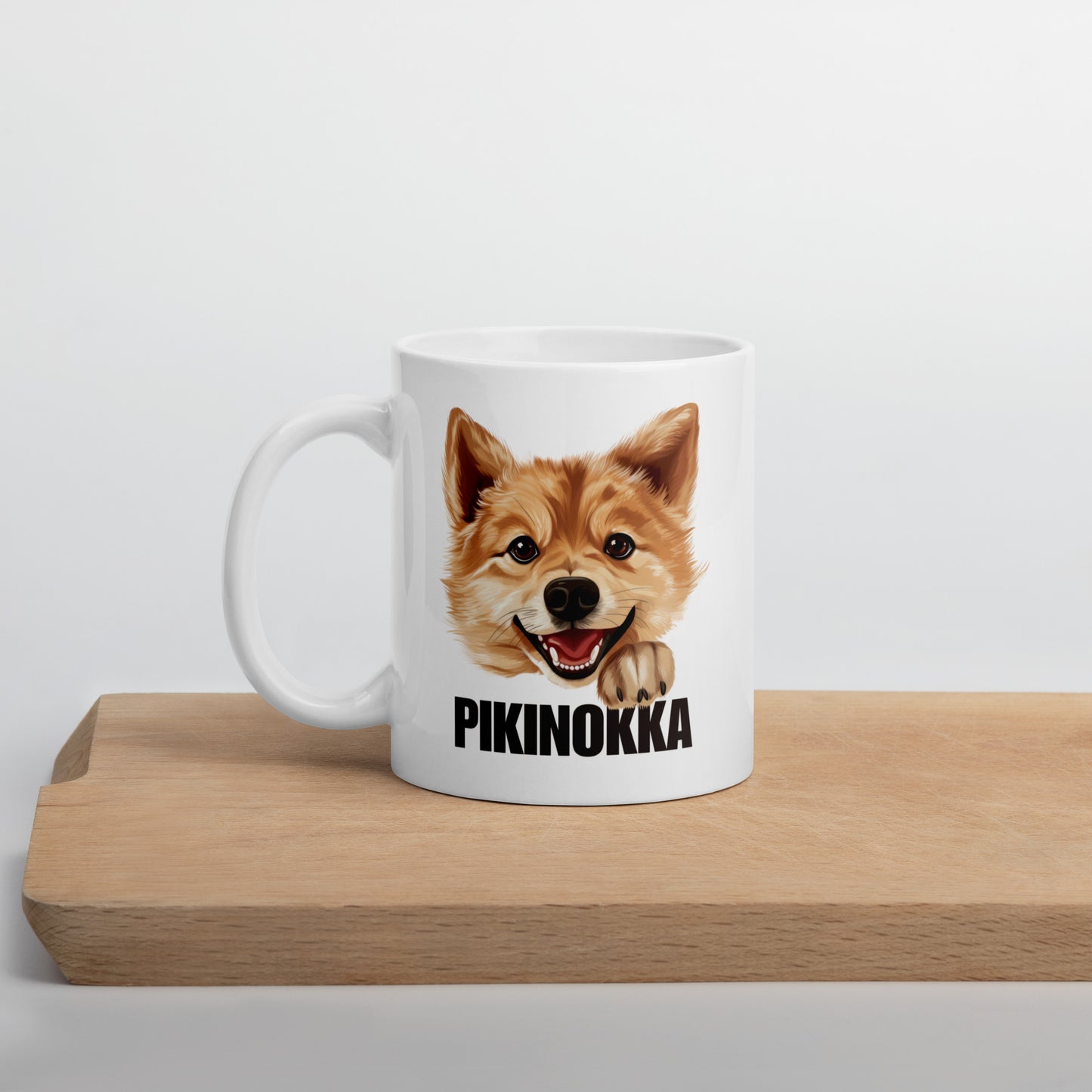 Suomenpystykorva muki, jossa koiran kuva ja teksti "pikinokka", kuvattuna leikkuulaudan päällä.