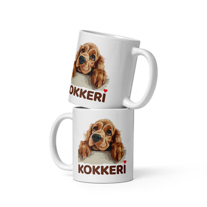 Kiiltävä valkoinen muki, jossa kurkkaavan cockerspanielin kuva ja teksti "Kokkeri" punaisella sydämellä.
